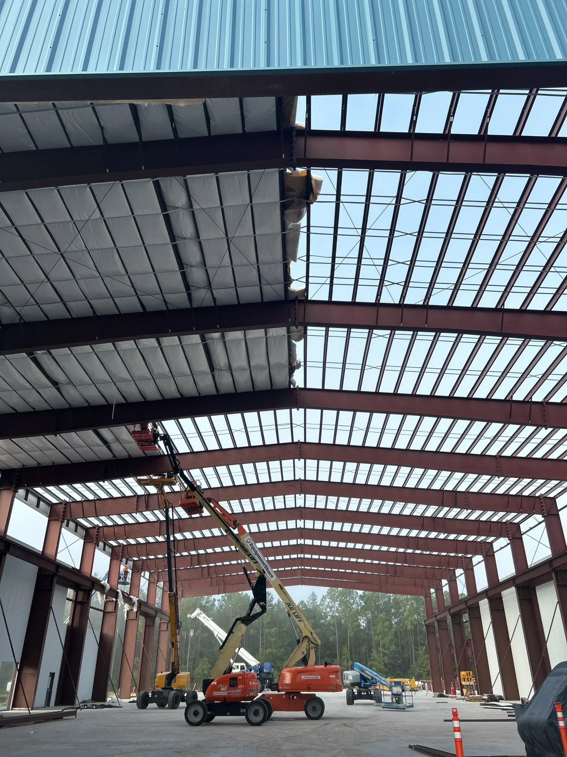 Construction workers using lifts to install a metal roof on a large industrial building with a steel framework and open sides.