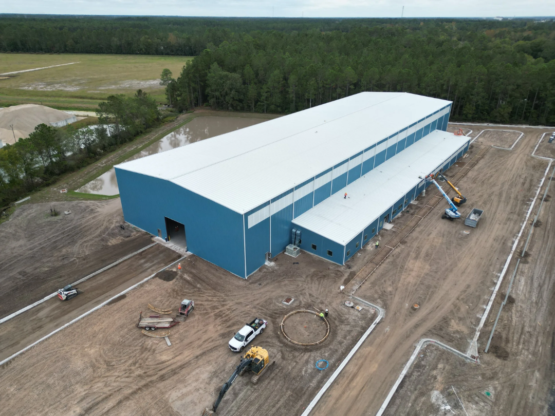 Aerial view of a large blue industrial warehouse under construction, with construction equipment, vehicles, and workers around the site, surrounded by open land and trees.
