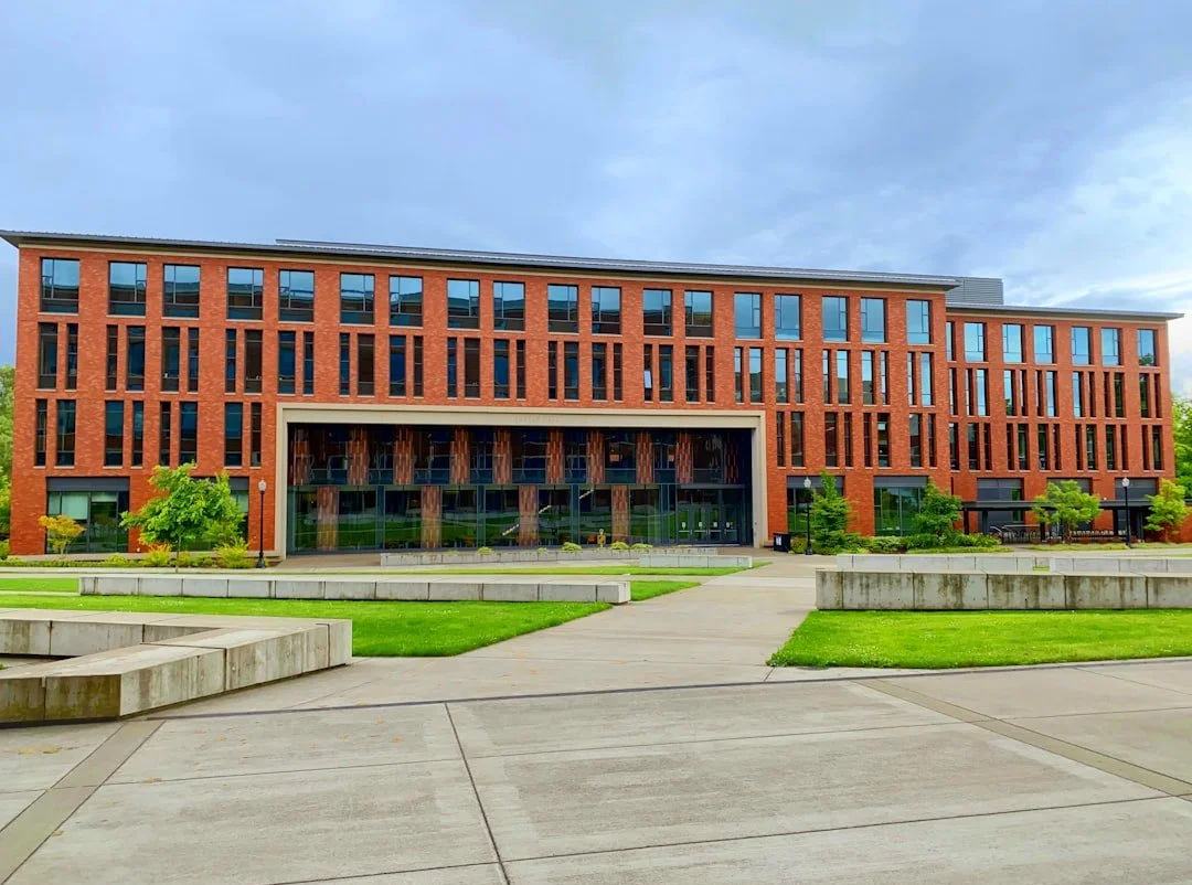A modern brick university building with multiple stories and large windows, set against a cloudy sky, surrounded by a paved walkway, green lawns, and small trees.