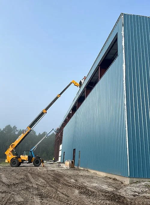 A construction site with a large blue warehouse and a crane lifting materials to the upper floor.